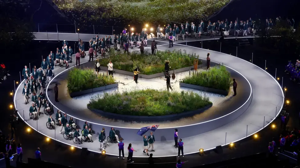 Australian athletes emerge on to the stage, with a number of Para-athletes at the front, some in wheelchairs, followed by the rest of the team. An Australian flag is waved at the front of the group.